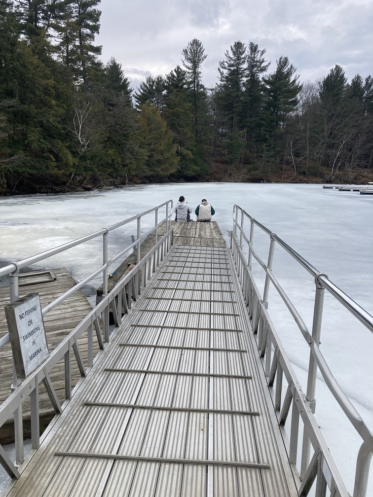 A dock juts out into the frozen ice of the marina at Keewaydin State Park in Alexandria Bay.  This photo of two people sitting at the end of the dock was taken in March