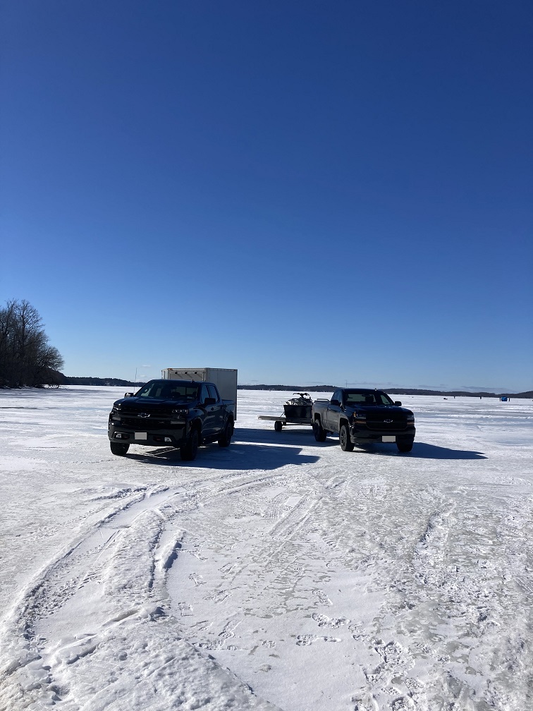 A bay in the St. Lawrence River is frozen in February, this photo shows two large pickup trucks with trailers parked on the ice