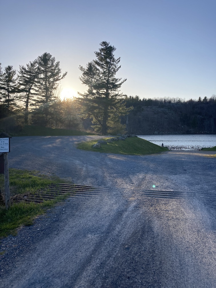 A photograph of the parking area at the boat launch site with the lake and the cliffs of the state forest in the background.  The sun is shining through the trees in the foreground